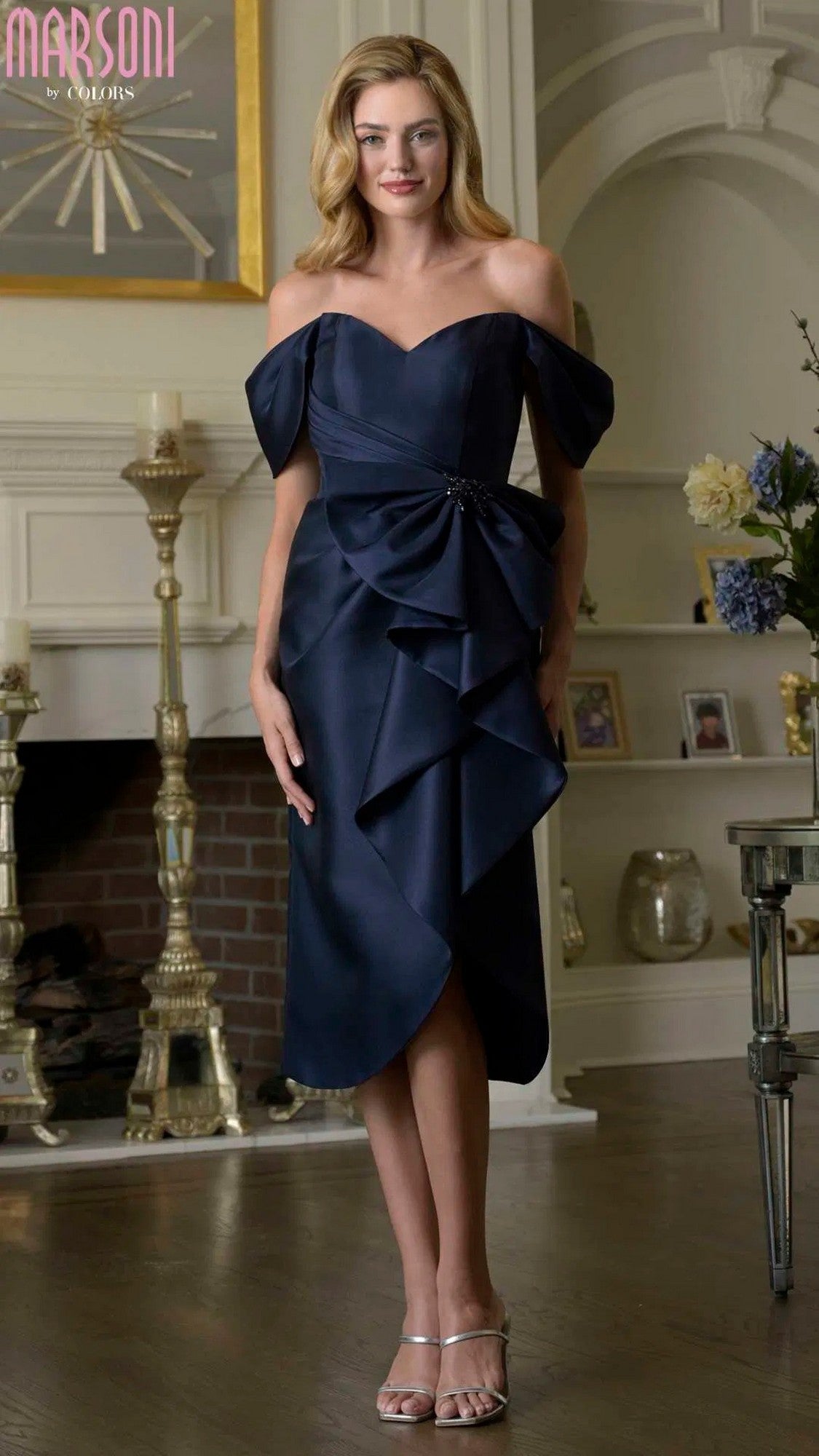 A woman stands indoors in front of a fireplace, wearing the Marsoni MV1389 knee-length semi-formal dress in navy with ruffle details and silver heels, surrounded by candlesticks and decorative items.